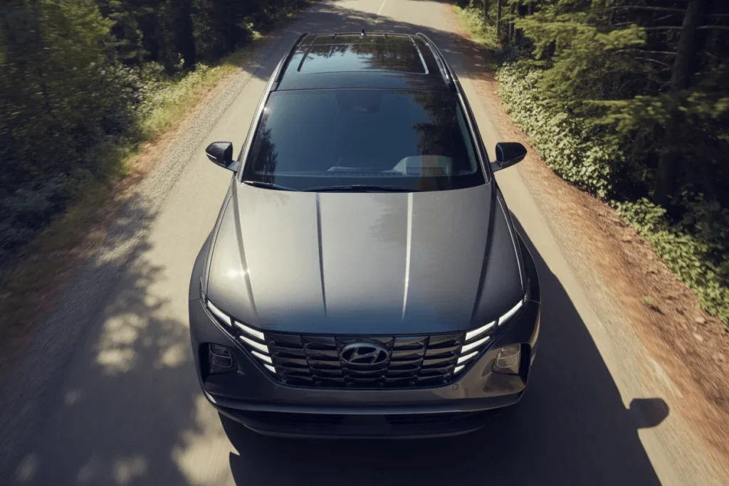 High front overhead drone view of 2025 Hyundai Tucson in Amazon Gray LED DRLs glowing on sunlit forest road High front overhead drone view of 2025 Hyundai Tucson in Amazon Gray LED DRLs glowing on sunlit forest road