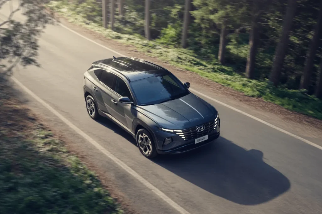 High front three quarter aerial of 2025 Hyundai Tucson cruising a sunlit pine road black roof LED DRLs alloys