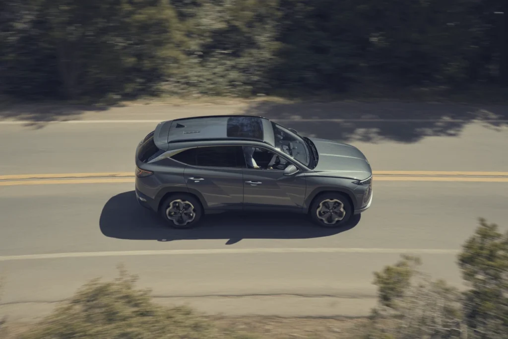 Overhead side profile of 2025 Hyundai Tucson cruising on two lane highway panoramic roof visible forest motion blur Overhead side profile of 2025 Hyundai Tucson cruising on two lane highway panoramic roof visible forest motion blur