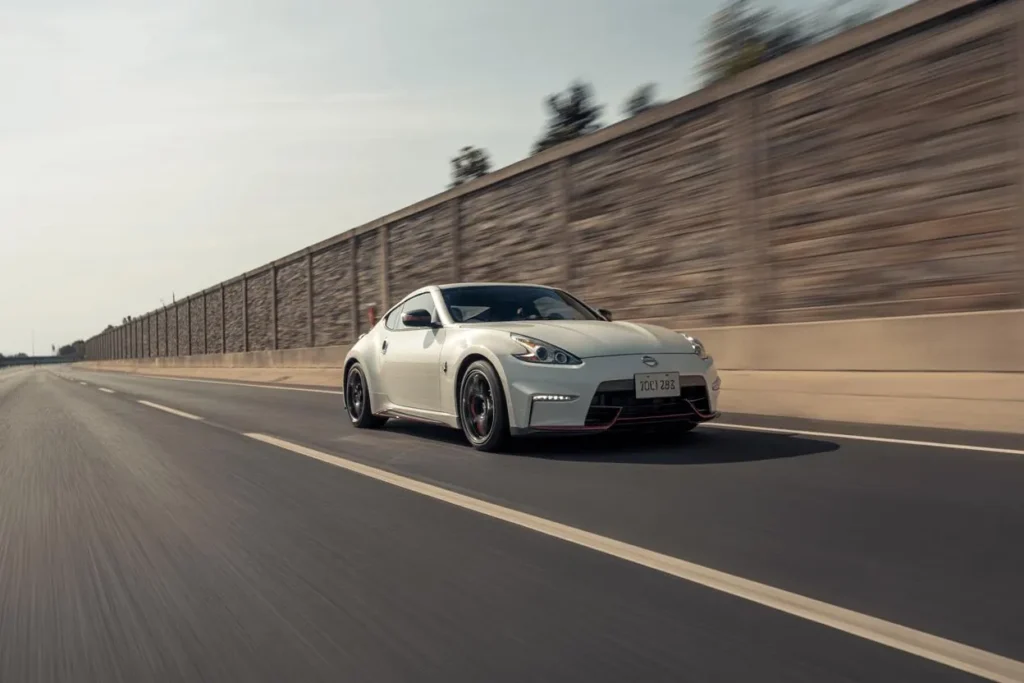 2014 Nissan 370Z Nismo front three quarter angle on sunny day pearl white sports car roadside sound wall black Nismo wheels 2014 Nissan 370Z Nismo front three quarter angle on sunny day pearl white sports car roadside sound wall black Nismo wheels