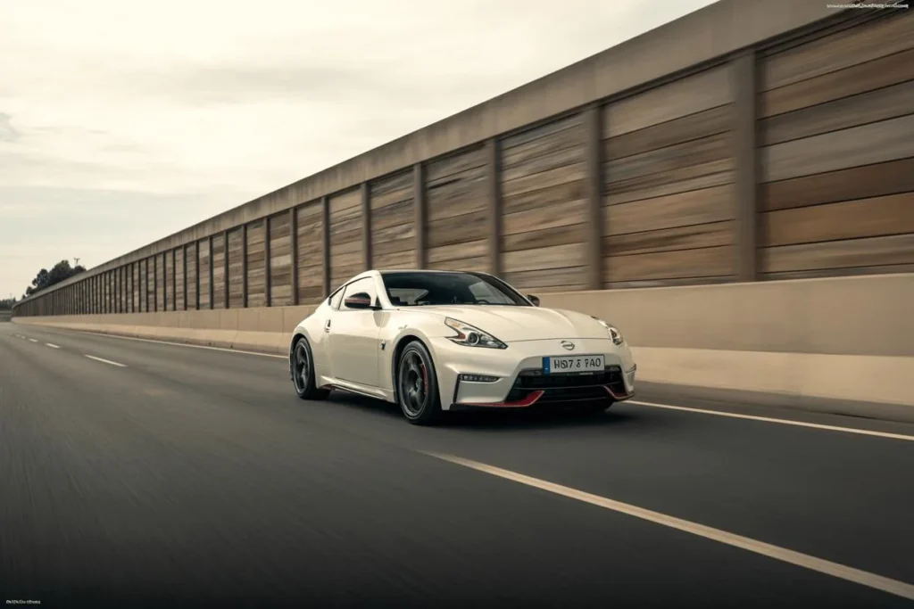 2014 Nissan 370Z Nismo front three quarter rolling shot beside long highway sound barrier pearl white sports coupe dark alloy wheels 2014 Nissan 370Z Nismo front three quarter rolling shot beside long highway sound barrier pearl white sports coupe dark alloy wheels