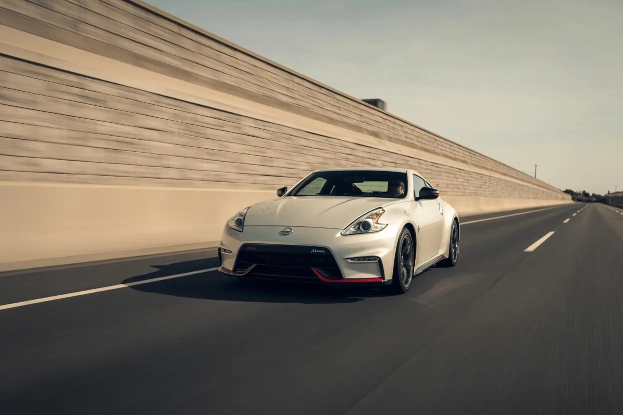 2014 Nissan 370Z Nismo front three-quarter shot on clear highway, pearl white bodywork, aggressive bumper, red lower splitter, motion blur.