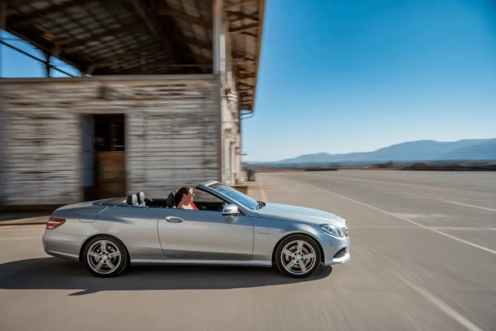 Side profile motion shot of 2014 Mercedes Benz E350 Cabriolet cruising past weathered building desert mountains blurred in background Side profile motion shot of 2014 Mercedes Benz E350 Cabriolet cruising past weathered building desert mountains blurred in background