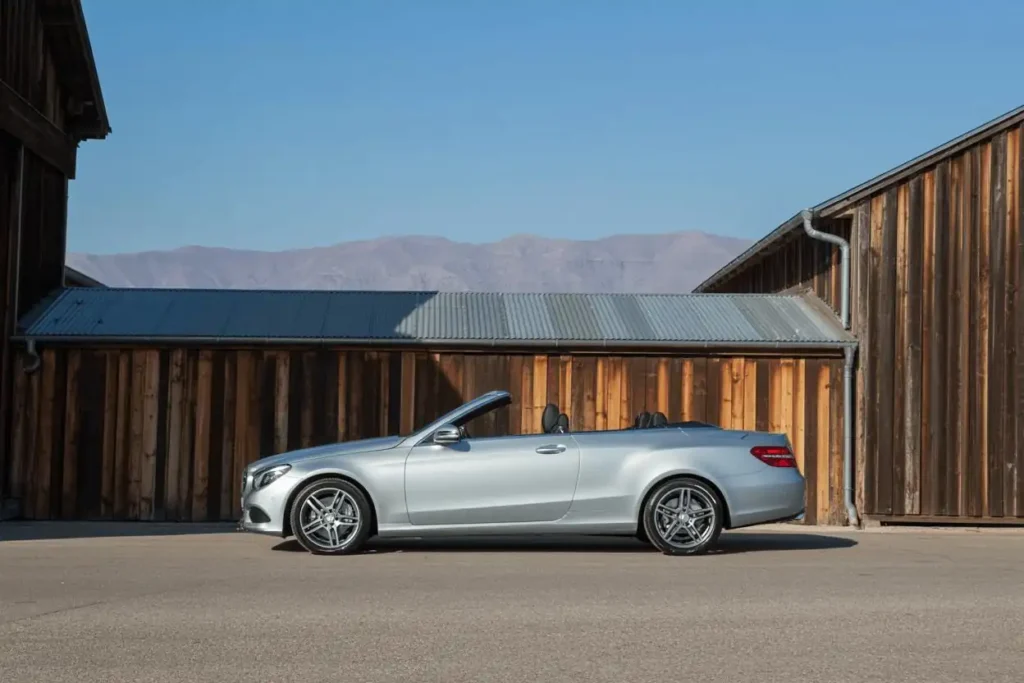 Side profile of 2014 Mercedes Benz E350 Cabriolet with roof down parked between wooden barns showcasing long body and alloy wheels Side profile of 2014 Mercedes Benz E350 Cabriolet with roof down parked between wooden barns showcasing long body and alloy wheels