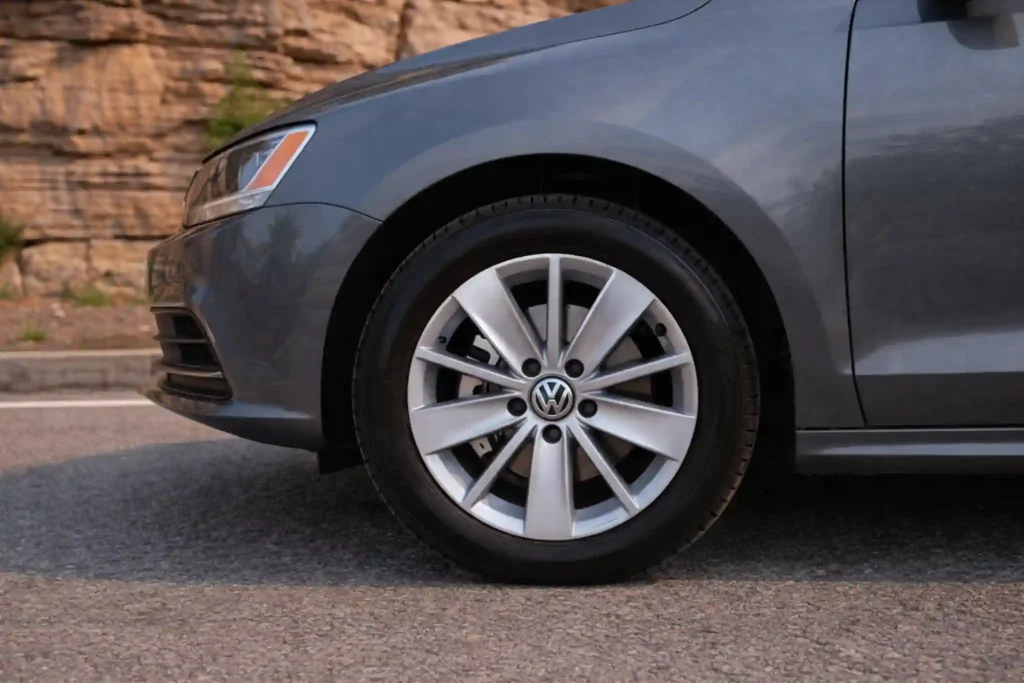 A gray 2015 Volkswagen Jetta 1.8T SE appears in a front left three quarter road shot with the grille headlights and front wheel visible against a rocky roadside backdrop A gray 2015 Volkswagen Jetta 1.8T SE appears in a front left three quarter road shot with the grille headlights and front wheel visible against a rocky roadside backdrop